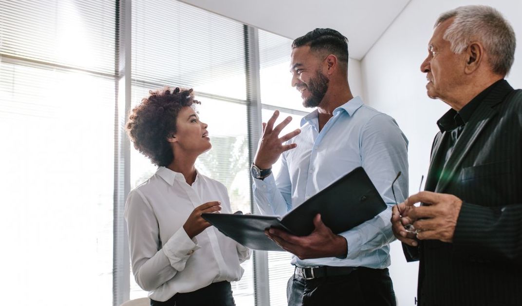 Three people talking while standing. One is holding a notebook as a reference.