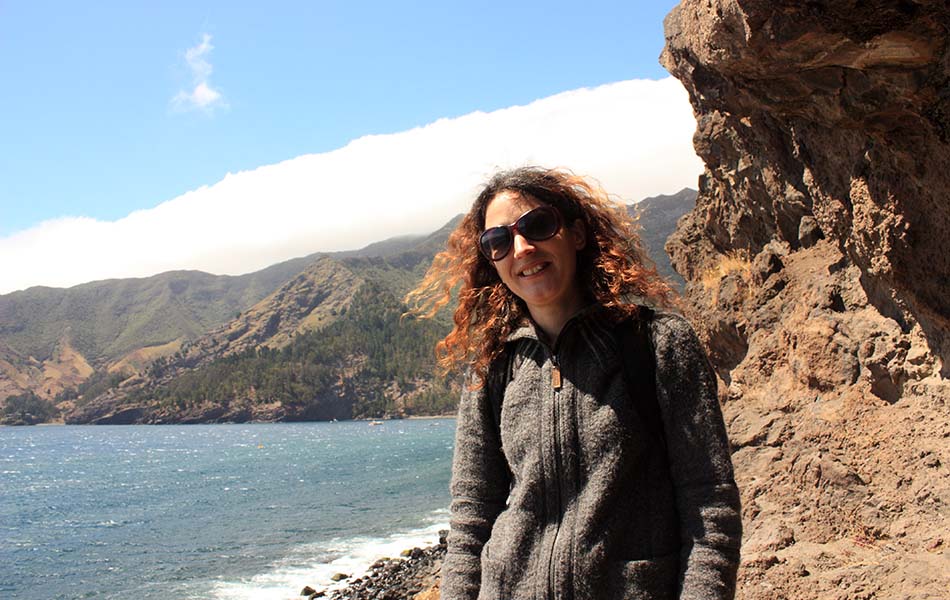 A photo of Dr. Dianne Newbury standing on the shores of Robinson Crusoe Island, with the ocean and part of the island in the background. She is wearing a grey jumper and sunglasses.