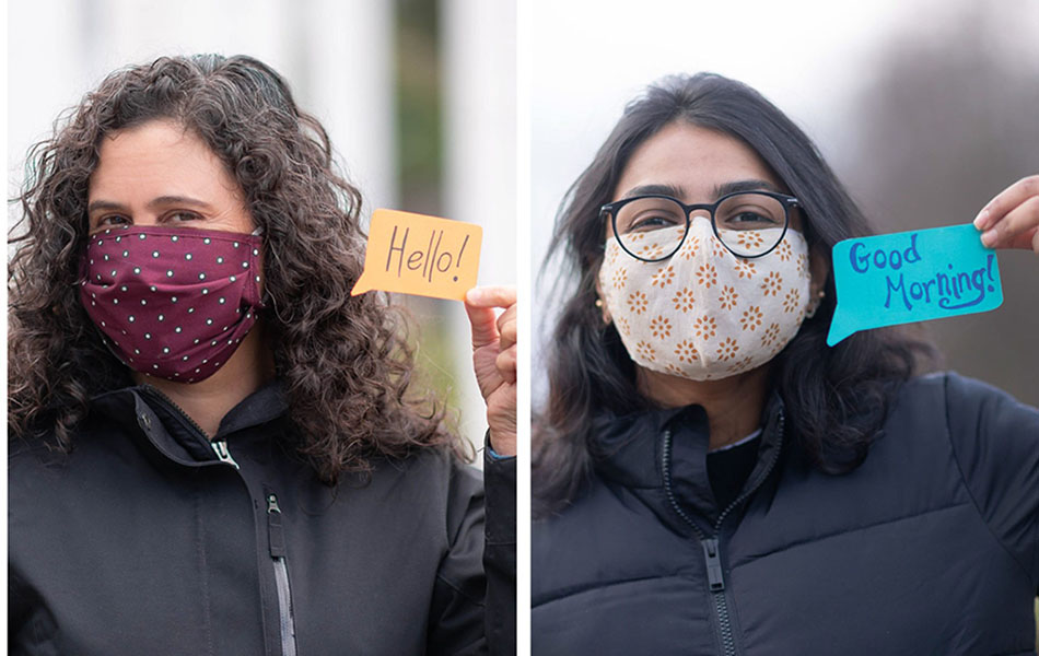 An image of two students wearing facemasks, and jackets, standing outside, holding up paper in the shape of speech bubbles with greetings written on the paper