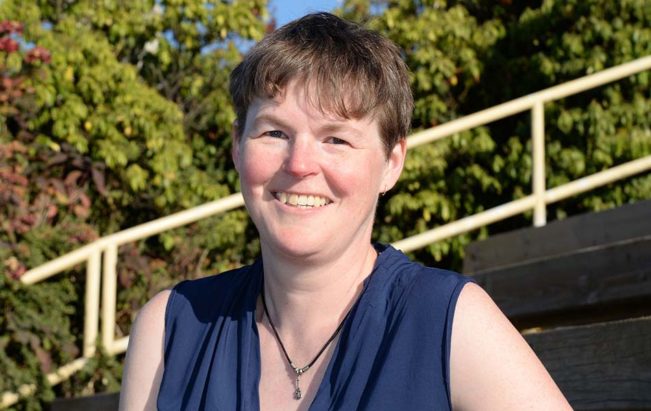 A headshot of Dr. Abel standing outside in front of trees and stone steps, wearing a blue top and smiling at the camera