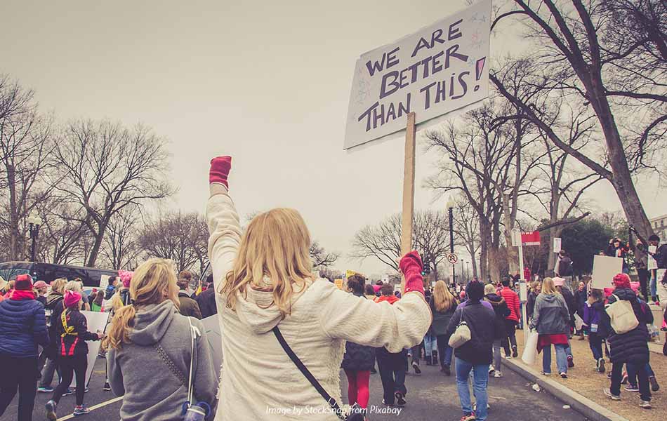 A photo from the back of protestors marching along a road, with a blonde woman in the foreground, her fist in a red glove and raised, holding a sign saying We are better than this