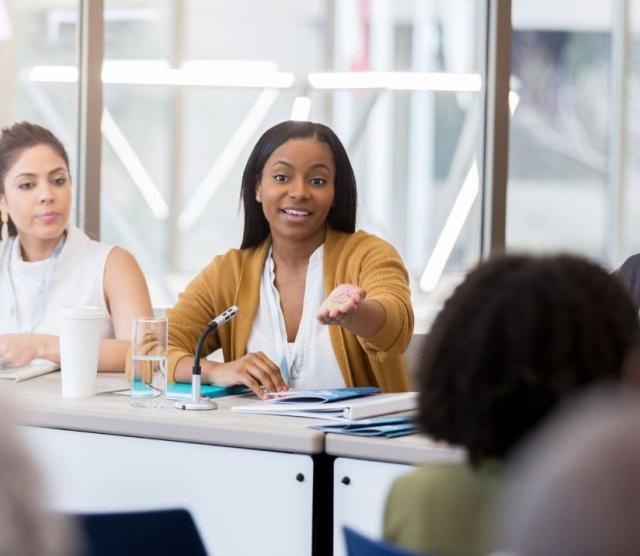 Three people sitting at a table, the person in the middle speaking with their hand out in gesture.