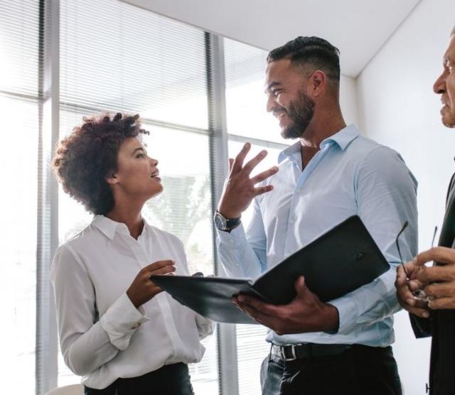 Three people talking while standing. One is holding a notebook as a reference.