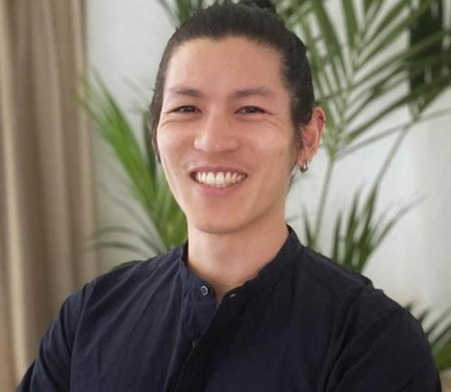 A headshot of Andrew Cheng standing with arms crossed, smiling at the camera, wearing a black shirt. Behind is palm plant and beige curtains