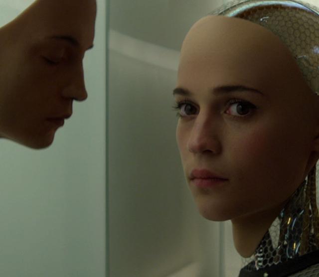 A still from the film of a human mask on a glass wall in front of which Ava the robot stands looking into the camera