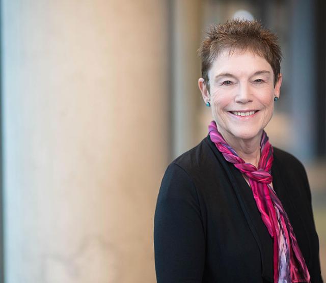 A photo of Bonny Norton smiling, standing beside a concrete pillar in a hallway. She is wearing a black jumper and a pink scarf