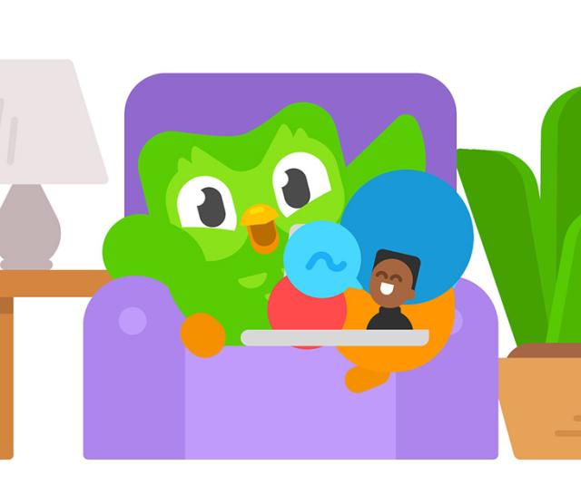 An image of the Duolingo owl siting in a purple armchair with a lamp and a potplant at the side and a laptop in its lap