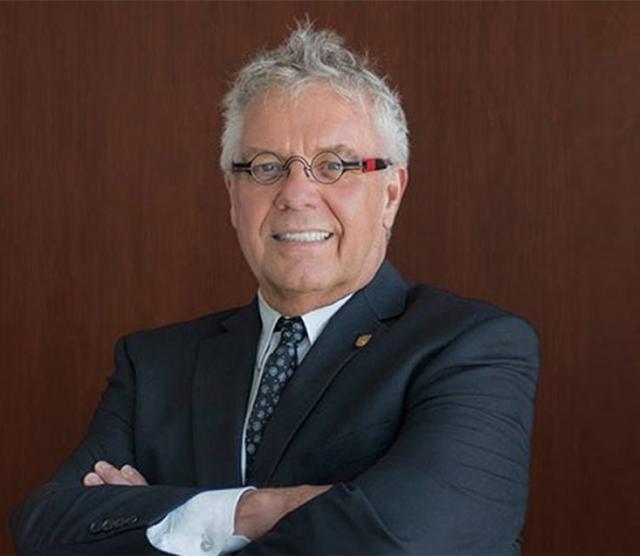 An image of Dean Blye Frank smiling, standing with arms crossed against a wooden panel wearing a suit and glasses