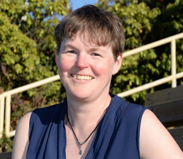 A headshot of Dr. Abel standing outside in front of trees and stone steps, wearing a blue top and smiling at the camera
