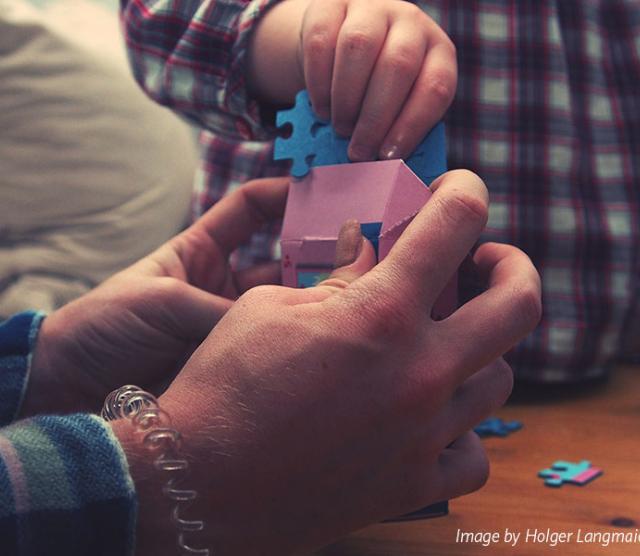 A close up on an adult's hands holding a pink box, with a child's hand placing a blue puzzle piece in the box