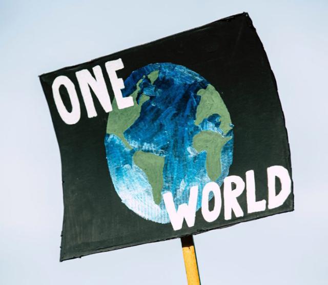 An image of a black poster held up in the sky which reads one world, with a picture of Earth