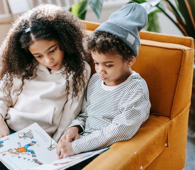 Two young children sitting together in an armchair, reading a book with pictures and text.