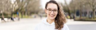 A headshot of Paris on UBC Main Mall, smiling, wearing glasses, and with her hair over one shoulder