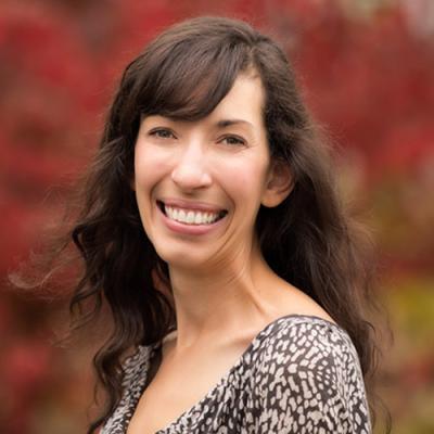 A headshot of Dr. Kathryn Accurso standing side on, smiling, in front of a tree with red leaves