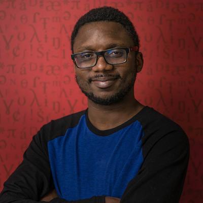 A headshot of Samuel Akinbo with arms folded, wearing glasses, standing against a red background