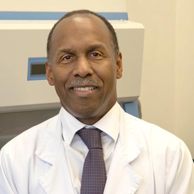 A headshot of Desmond Nunez smiling, sitting in a clinic room, wearing a tie and a lab coat