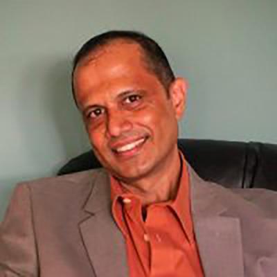 A headshot of Reginald D'Silva smiling, and wearing an orange shirt under a tan jacket, sitting in a chair against a green wall