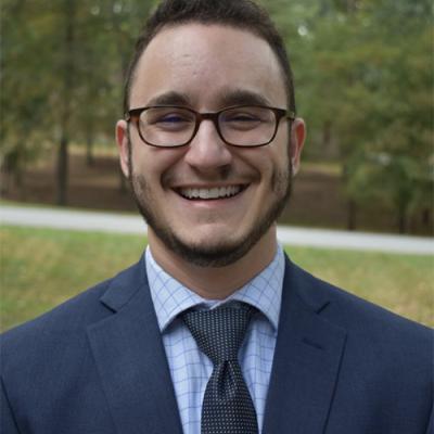 A headshot of Ethan Kutlu smiling, wearing glasses and a suit and tie, with green grass and trees in the background