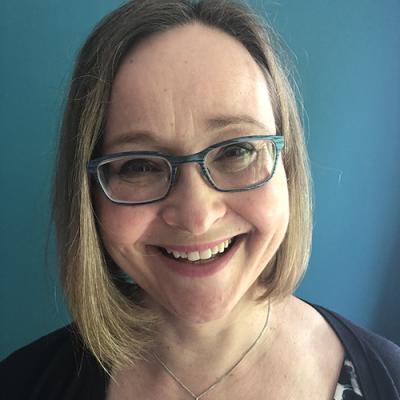 A headshot of Daniela O’Neill smiling at the camera and wearing glasses, while she stands against a blue background