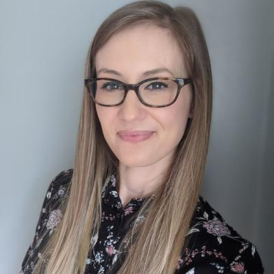 A headshot of Sara Van Dan Acker smiling at the camera wearing a patterned shirt, glasses, and standing against a grey wall.