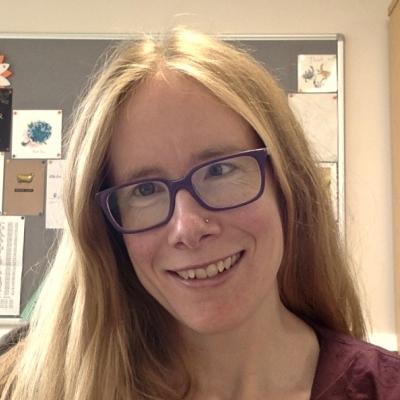 A headshot of Jenny Thomson smiling. Dr Thomson is wearing glasses and has long hair.