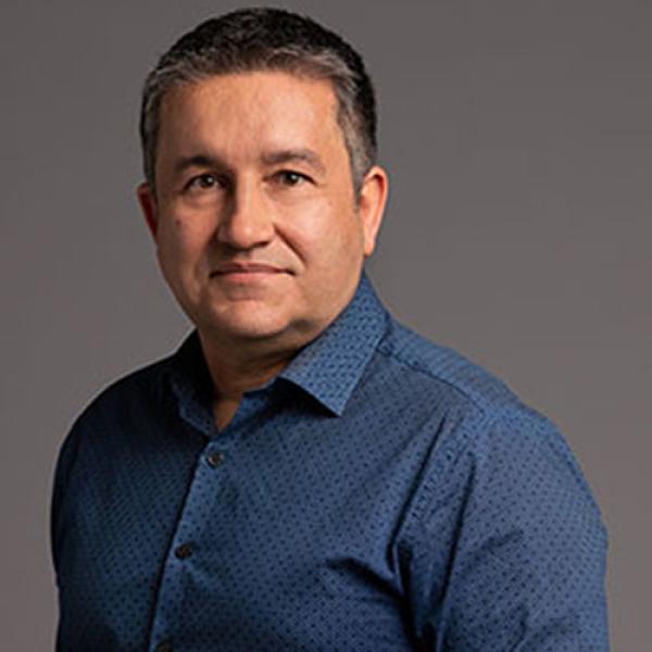 A headshot of Alfred Hermida wearing a blue shirt and standing in front of a grey background