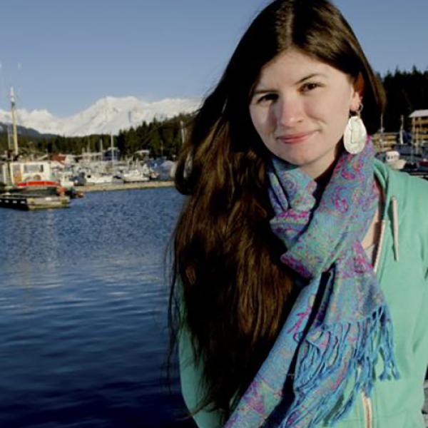 An image of Heather Burge standing in front of a harbour, wearing a green jumper and multicoloured scarf