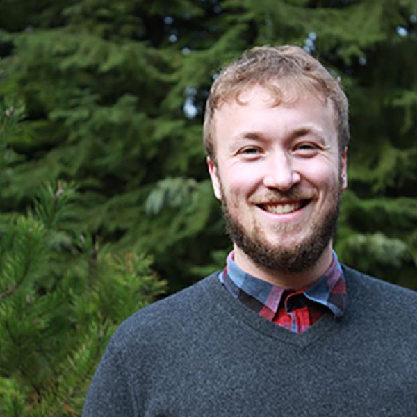 A headshot of Aidan Pine, smiling, standing in front of a tree.